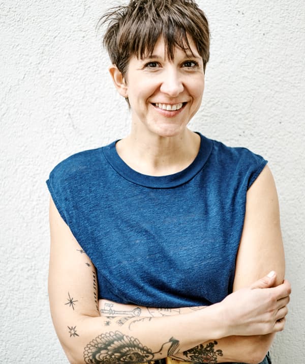 Una donna, con i capelli corti e tatuaggi sulle braccia, indossa una maglia blu senza maniche, appoggiata contro una parete bianca.