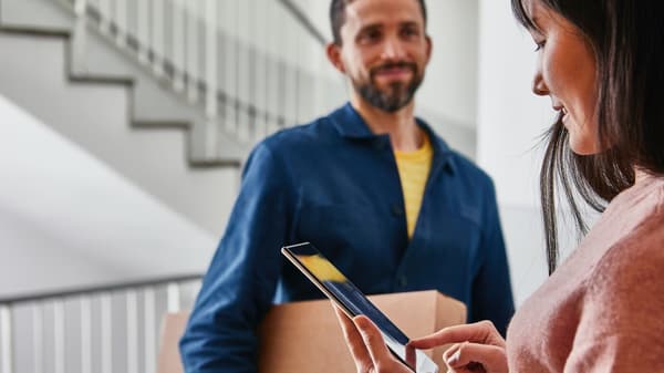 Una donna controlla sullo smartphone i dettagli della consegna del suo pacco mentre l’addetto al recapito attende sorridente con il pacco.