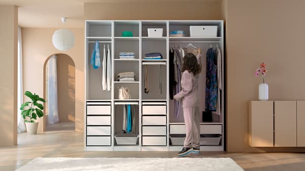 Una donna dai capelli scuri con un tailleur pantalone rosa appende un vestito a un guardaroba PAX bianco.