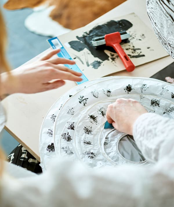 Una donna decora con foglie di felce un paralume bianco in carta ancora piegato, utilizzando un timbro intinto nella tempera nera.