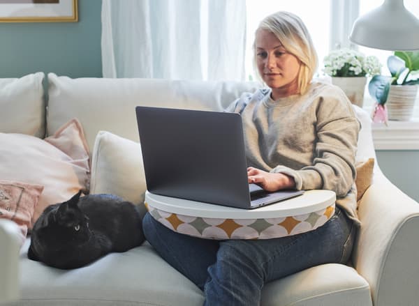 Una donna seduta su un divano bianco accanto af un gatto. Ha un supporto per pc e sta lavorando.
