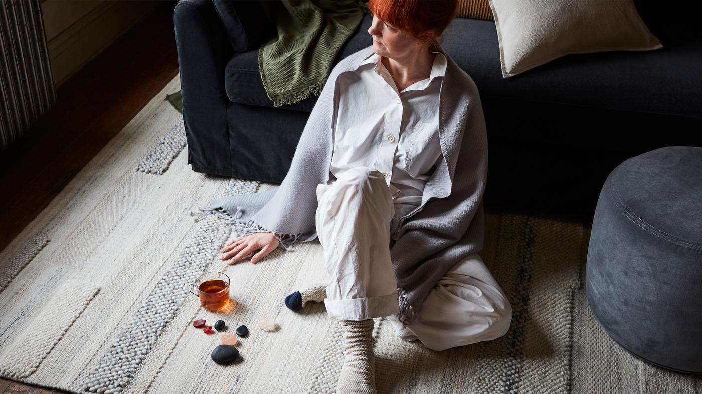 Una donna seduta su un tappeto BRÖNDEN beige tessuto a mano, accanto a un poggiapiedi e a un divano FÄRLÖV grigio scuro con cuscini e plaid.