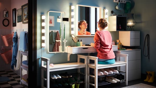 Una donna, seduta su una panca con vano per scarpe, si trucca nell’ingresso trasformato in angolo make-up utilizzando una mensola con cassetti, luci e specchi - IKEA