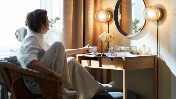 Una donna si rilassa su una poltrona e prende una tazza da una toeletta con sopra una rivista, un vaso di fiori e una scatola trasparente - IKEA