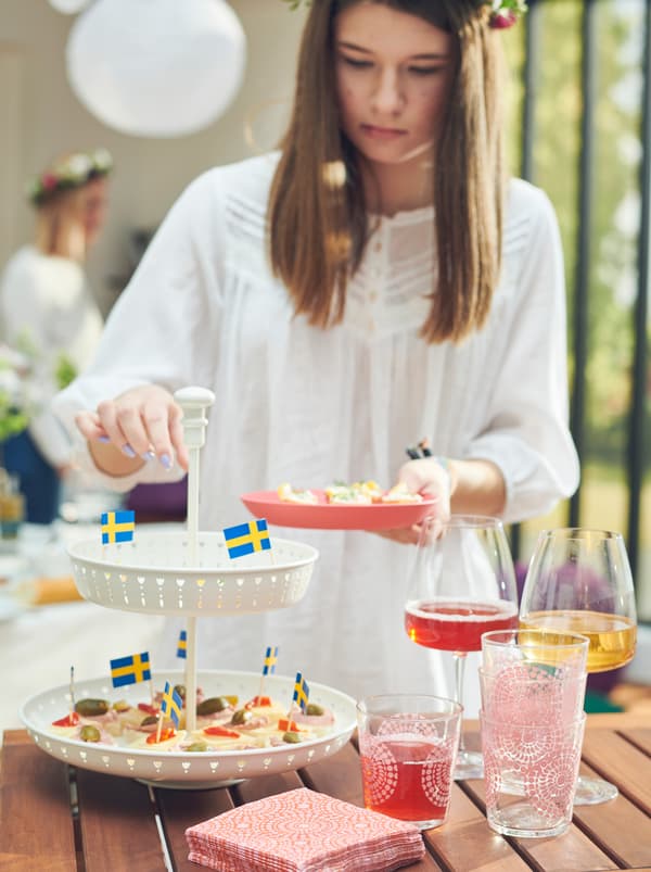 Una donna si serve degli stuzzichini da un’alzata GARNERA appoggiata su un tavolo, accanto a dei bicchieri da vino rosso DYRGRIP - IKEA