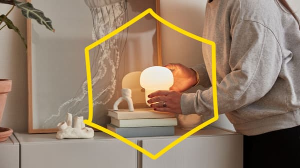 Una donna tiene in mano la lampada da tavolo TOKABO sopra una pila di libri su un mobile bianco, dietro ai libri è appesa una foto incorniciata di una medusa.