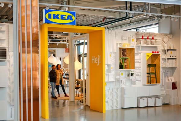 Una entrada amarilla brillante con el logo azul y amarillo de IKEA da la bienvenida a los visitantes a una tienda, con dos personas mirando en el interior. El área circundante está decorada con diversas soluciones de almacenamiento y muebles blancos y neutros, típicos del estilo de exposición de IKEA.