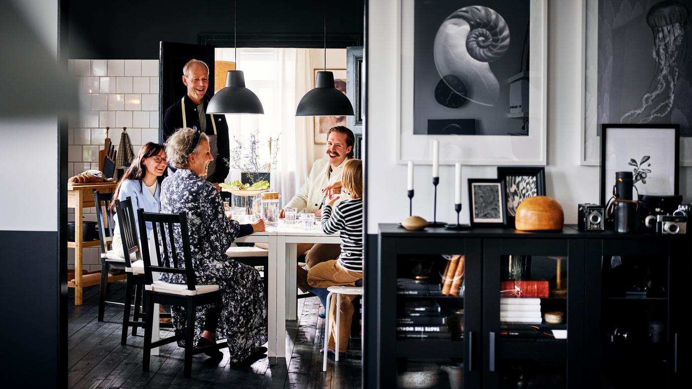 Una famiglia sorridente seduta su sedie STEFAN marrone-nero intorno a due tavoli MELLTORP bianchi in una zona pranzo in bianco e nero - IKEA