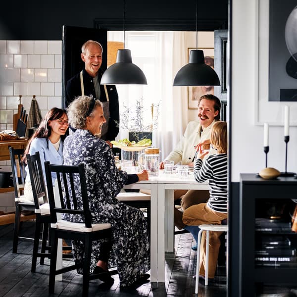Una familia sonriente sentada en sillas negras-café STEFAN alrededor de dos mesas blancas MELLTORP en un comedor blanco y negro.