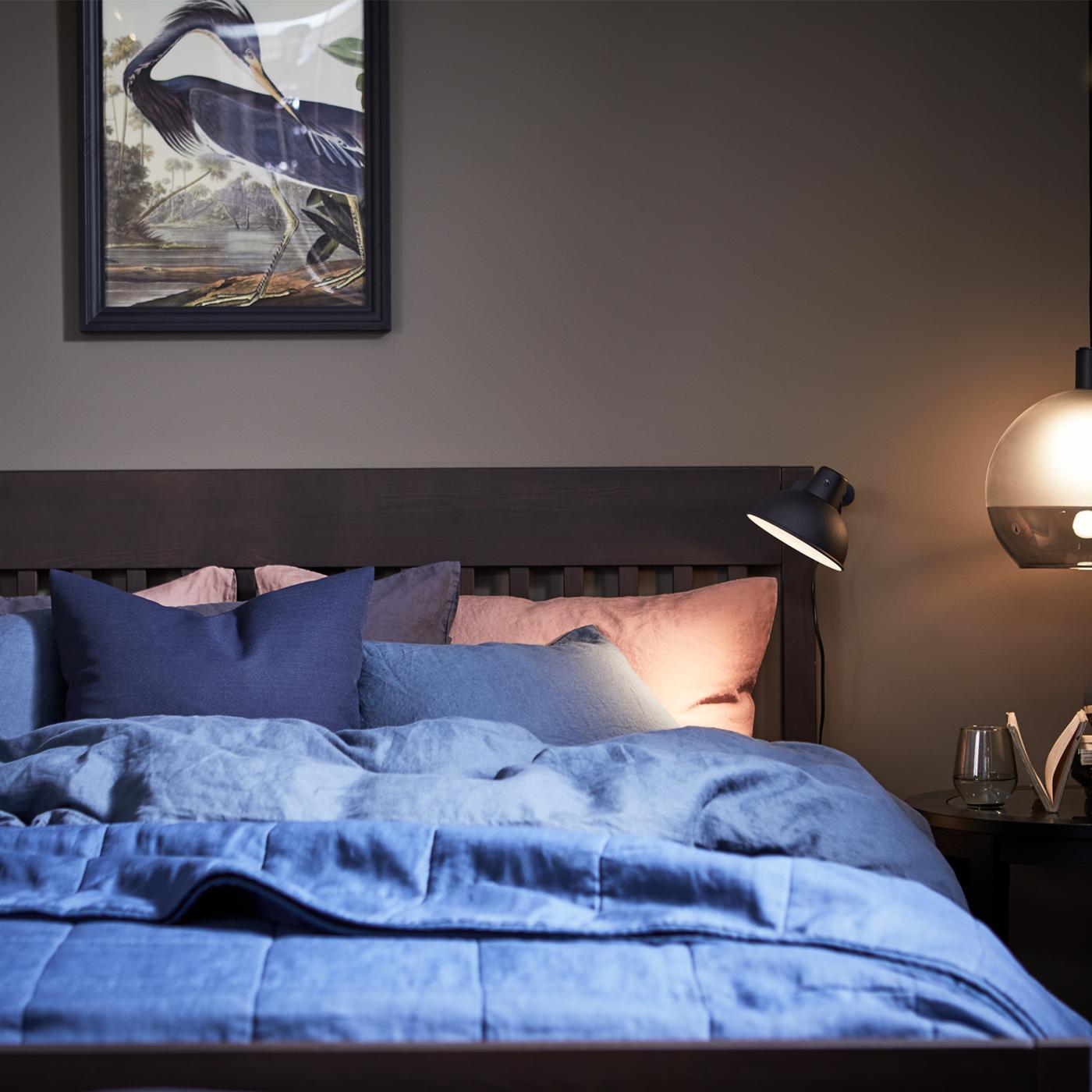 Una funda nórdica PUDERVIVA de color azul oscuro sobre una base de cama IDANÄS con almacenamiento y una pantalla colgante JAKOBSBYN encendida a la derecha..