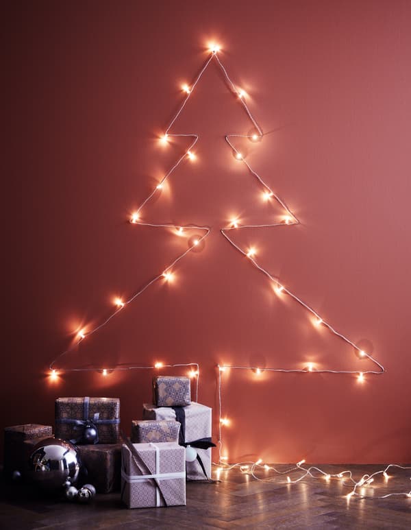 Una garlanda lluminosa en una paret amb forma d’arbre de Nadal, amb claus com a suport i amb regals al terra.