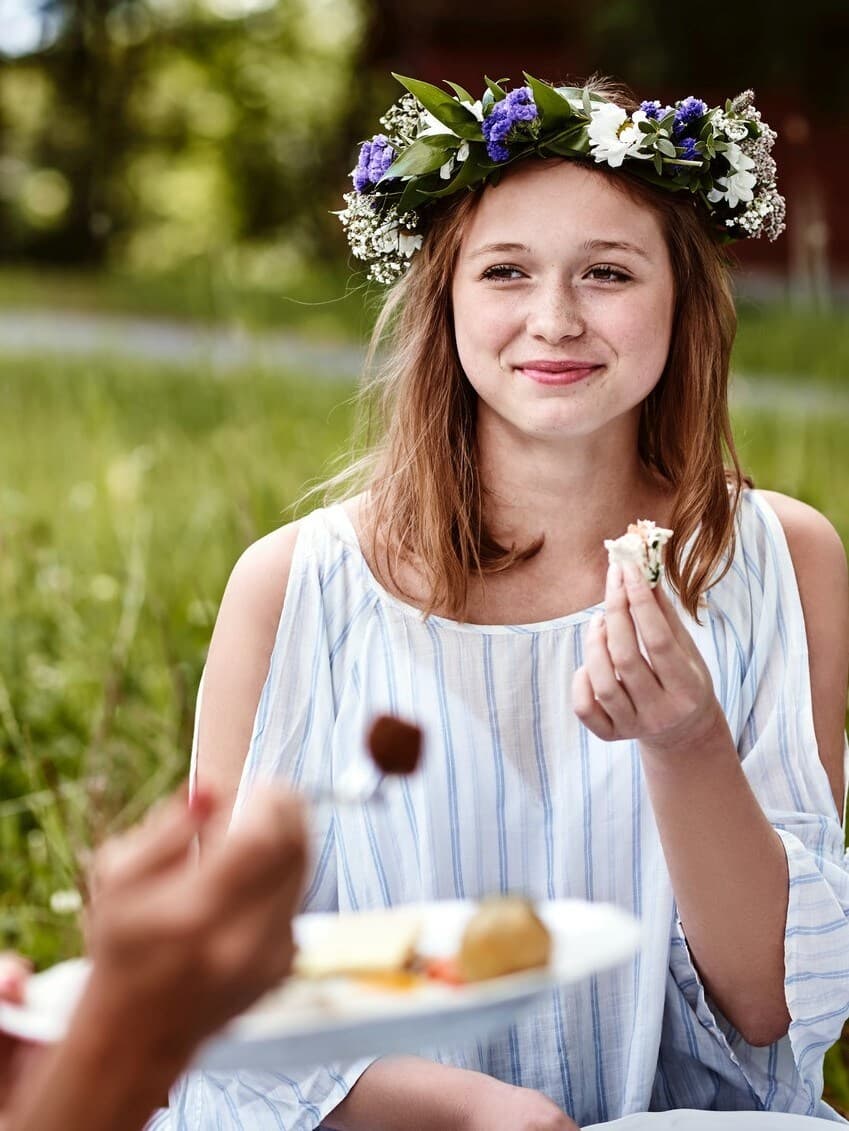 Una giovane ragazza con una coroncina di fiori in testa mentre sta mangiando in un prato in occasione della festa del Midsommar..