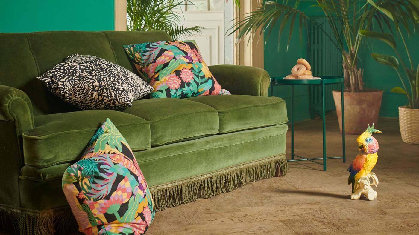 Una grande stanza con varie piante e un divano in velluto verde su cui sono sistemati dei cuscini con fodere GRIMHILD e NÄBBFLY con motivi esotici - IKEA