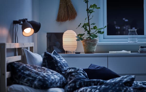 Una habitació a les fosques amb roba de llit de color blau, lleugerament il·luminada amb un llum de paper decoratiu i un llum de taula.