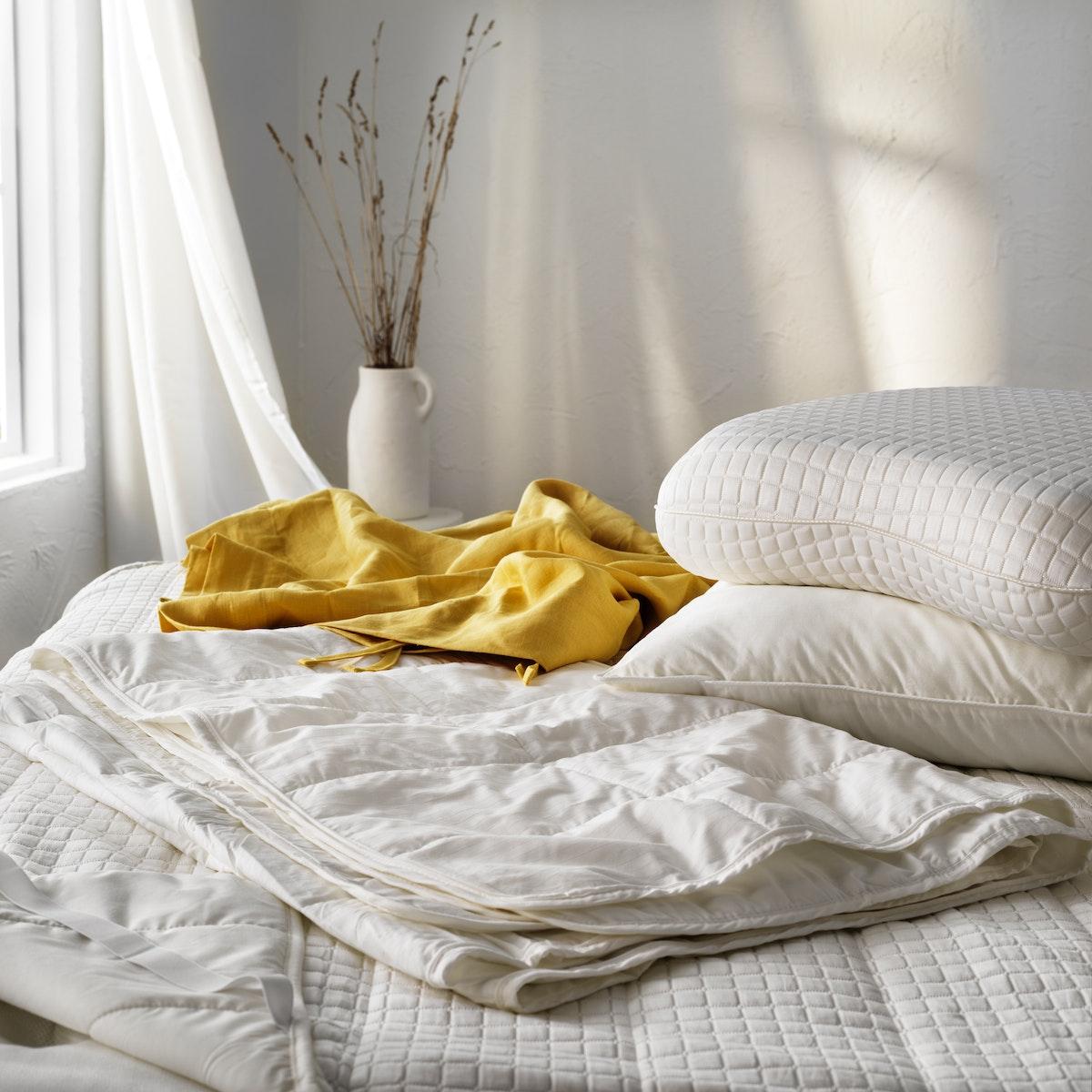 Una habitación blanca, en donde se muestra una cama con ropa de cama ligera, un arreglo floral y una manta amarrilla.