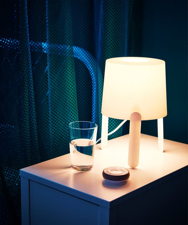 Una lampada accesa sul comodino con accanto un bicchieri e un caricabatterie wireless.