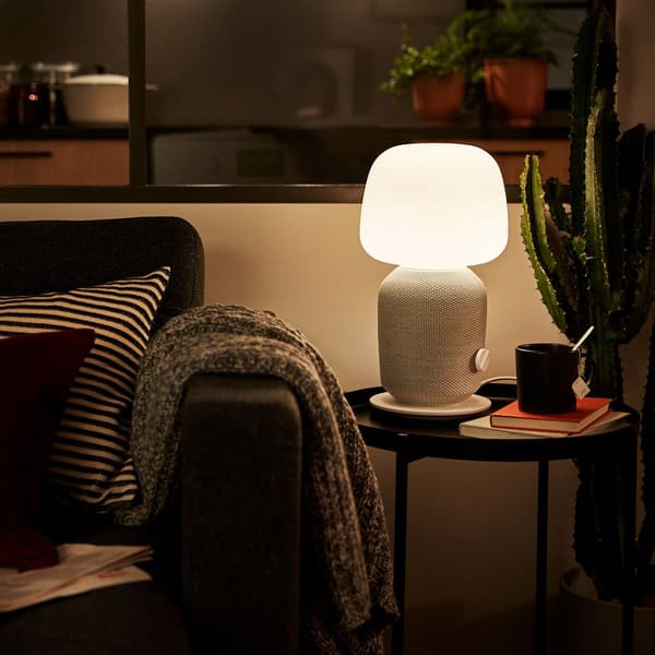 Una lampada con cassa accessa su un tavolino  GLADOM. Accanto si vede un divano con un plaid e cuscini.