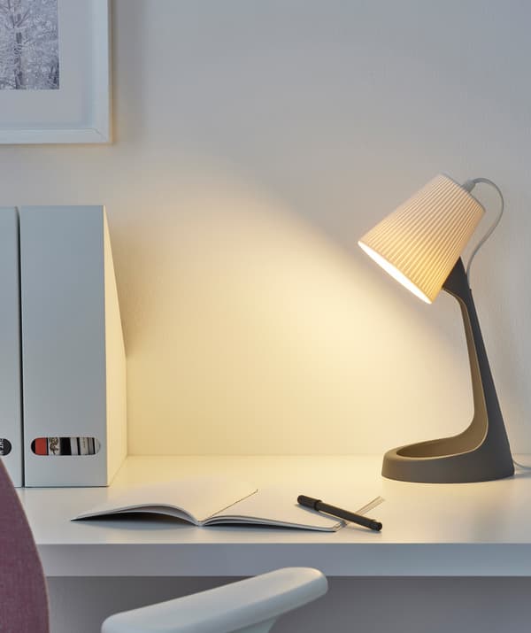 Una lampada da lavoro con base grigia e paralume bianco e un quaderno su una scrivania bianca.