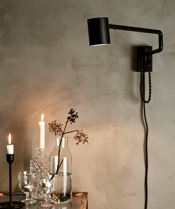 Una lampada da parete nera su una parete grigia illumina un ripiano in vetro con bicchieri, candele e candeline.