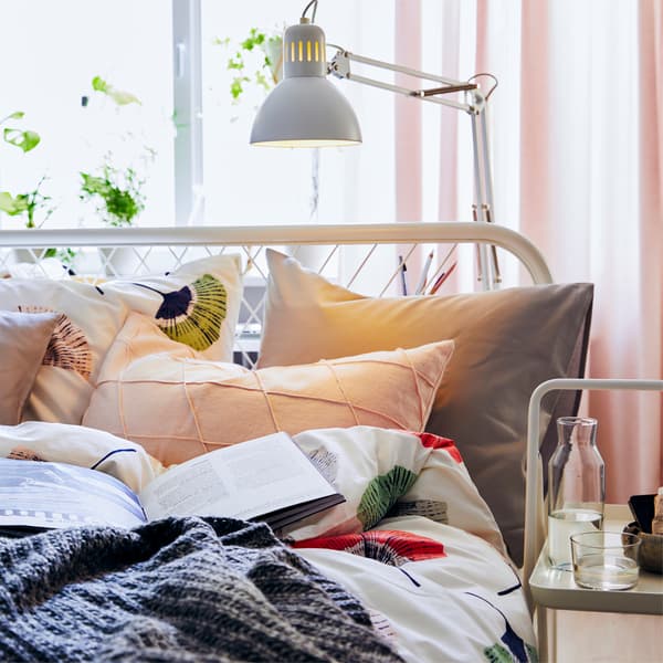 Una lampada da tavolo illumina un libro e dei cuscini su un letto - IKEA
