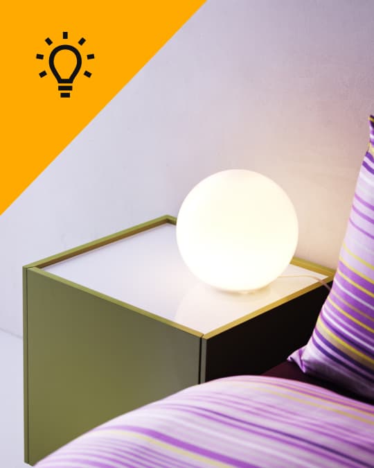 Una lámpara de mesa FADO encendida al costado de una cama sobre un velador.
