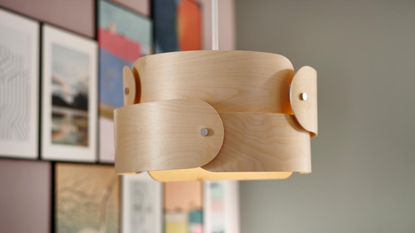 Una lámpara de techo de abedul SÖDÅKRA, con capas de madera superpuestas, colgada junto a una pared colorida con cuadros.