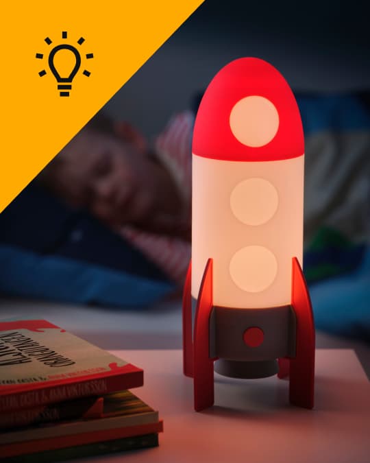 Una lámpara infantil de cohete encendida en un velador al costado de la cama de un niño.