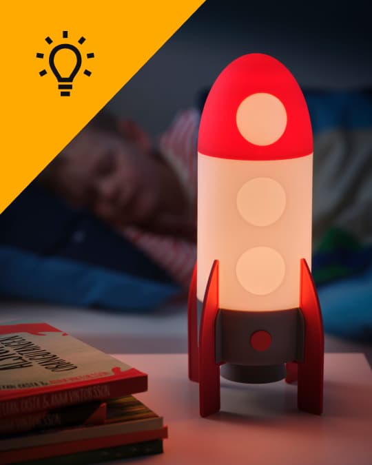 Una lámpara infantil IKEA con forma de cohete encendida sobre un velador de un dormitorio infantil mientras un niño duerme en segundo plano.
