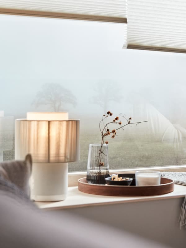 Una lámpara SYMFONISK  de altavoz con pantalla textil en el alféizar de una ventana. Fuera hay un árbol, apenas visible a través de la niebla.