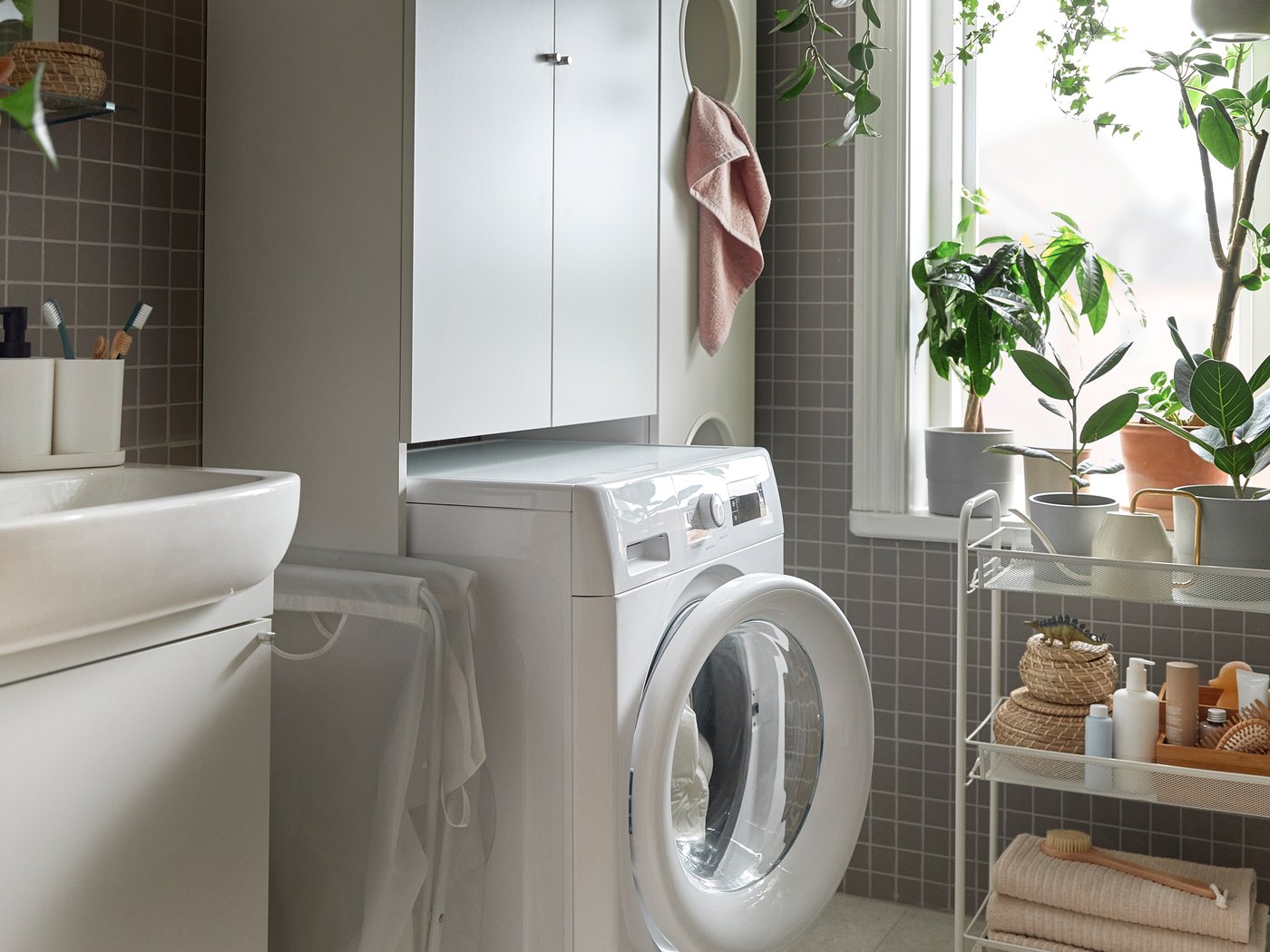 Una lavatrice bianca in un bagno, all'interno di una soluzione per lavanderia ENHET. Sulla finestra ci sono delle piante e accanto c'è un carrello - IKEA.