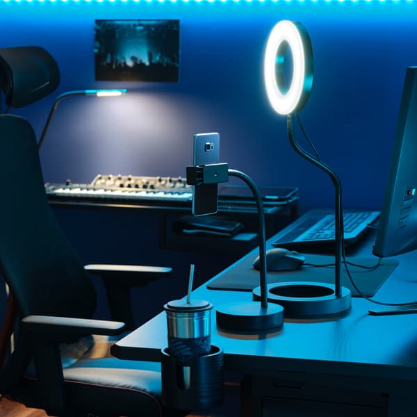 Una luce ad anello LÅNESPELARE e un supporto per telefono su una scrivania e, davanti, una sedia da gaming MATCHSPEL in una stanza da gaming con illuminazione blu.