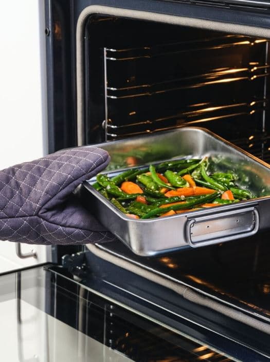 Una mà amb un guant de forn col·loca una safata metàl·lica amb verdures a l’interior d’un forn calent.