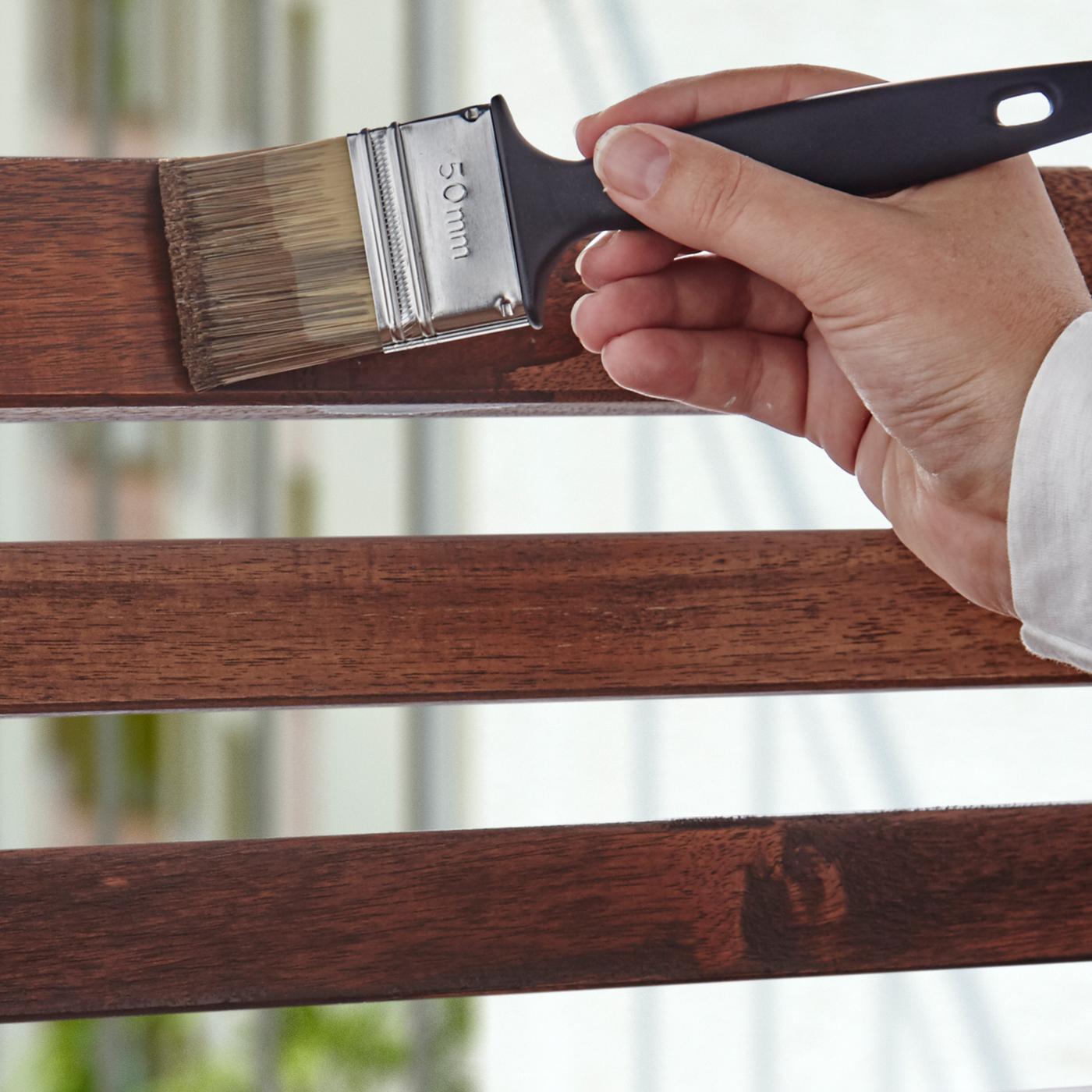 Una mà amb una brotxa amb pintura negra aplica vernís a la superfície d'un moble d'exterior.