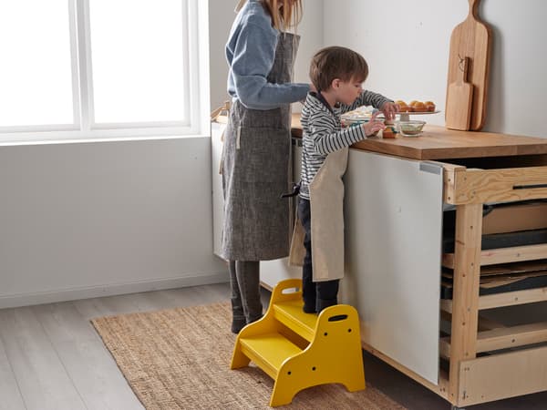 Una madre y su hijo cocinando, ambos tienen delantales de cocina. El niño está parado sobre un piso escalera amarillo.