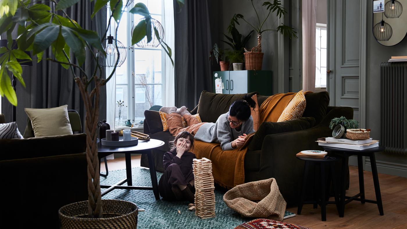 Una mamma distesa su un divano FÄRLÖV gioca con la sua bambina seduta sul pavimento. Lampade a sospensione GRINDFALLET sul soffitto.