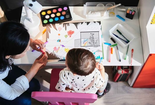Una mamma e un bambino seduti a una scrivania mentre stanno colorando. Il bambino è seduto su una sedia MAMMMUT e sulla tavola ci sono acquerelli, fogli, biro colorate, pennelli e altri oggetti.