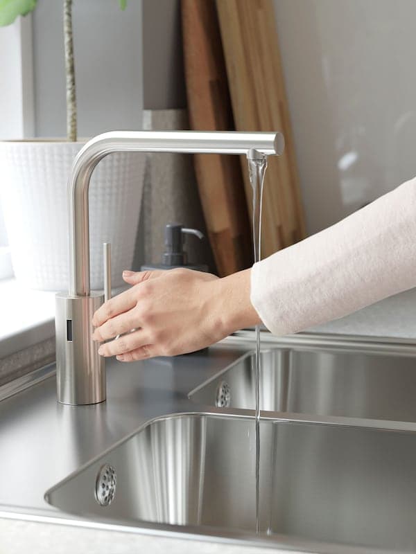Una mano ajusta el flujo de agua de un grifo de cocina GLYPEN en acero inoxidable junto a un cepillo para lavar platos TÅRTSMET.
