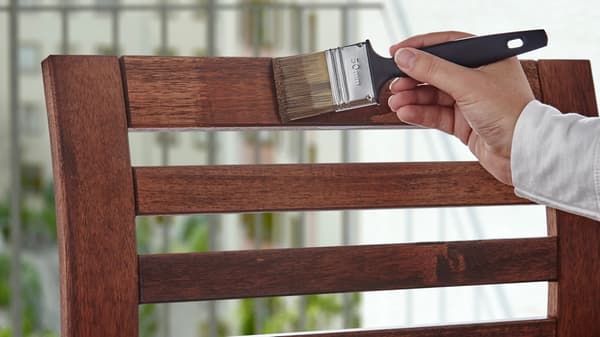 Una mano con un pennello, che vernicia con mordente lo schienale di una sedia da esterno in legno.