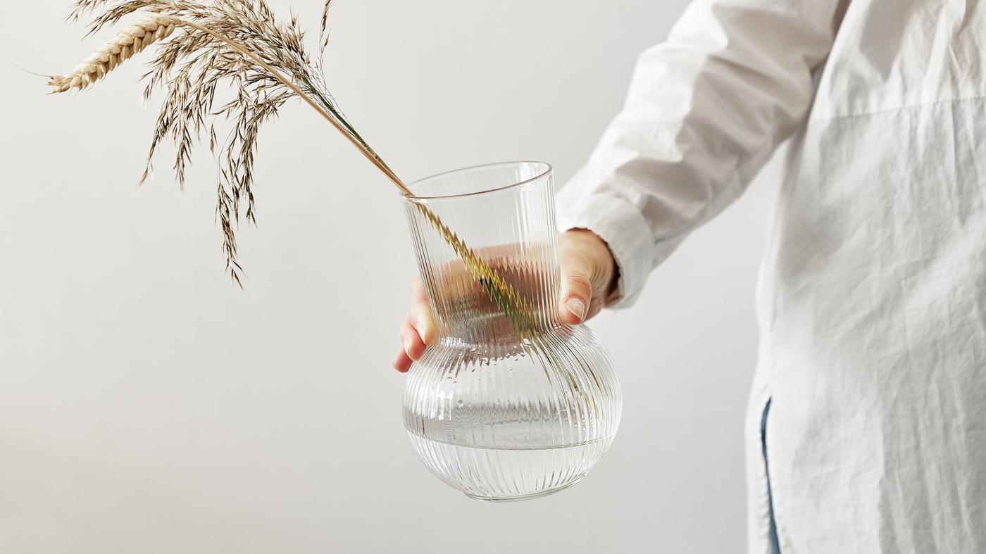 Una mano estirada sostiene un jarrón PÅDRAG de vidrio transparente con trigo y ramitas secas.