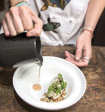 Una mano mentre completa un piatto bianco con gelato fatto con SILL MATJES aringhe, burro di frutta secca selvatica mista, foglie di aegopodium podagraria, pane al lichene islandico essiccato e sbriciolato e terra di radici fermentate.