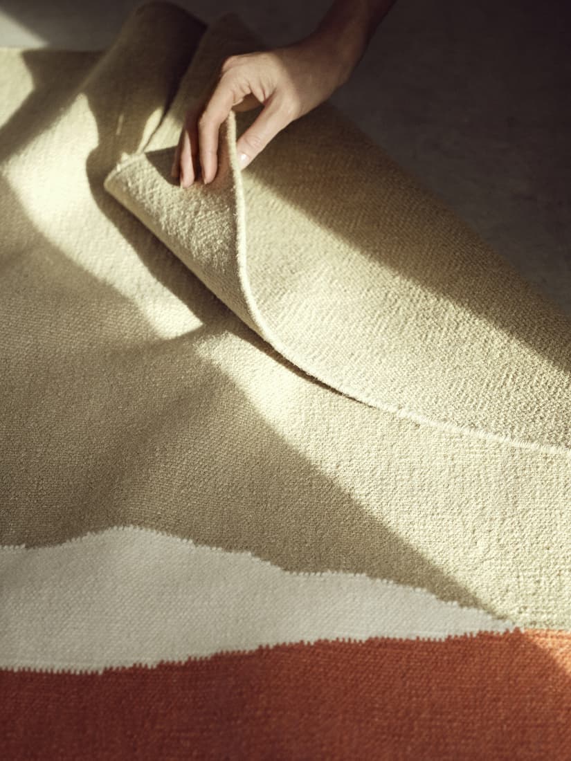 Una mano sosteniendo una alfombra y debajo de esa alfombra hay otra alfombra con tonos de color tierra.