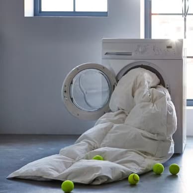 Una máquina de lavar centrifuga con un edredón que sobresale por la puerta, se ven unas pelotas de tenis esparcidas por el piso.