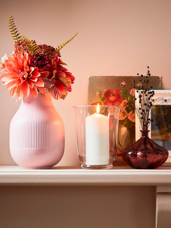Una mensola in legno bianco con un vaso di fiori rosa GRADVIS. Accanto, una candela accesa in un contenitore in vetro POMP e un altro vaso di fiori.