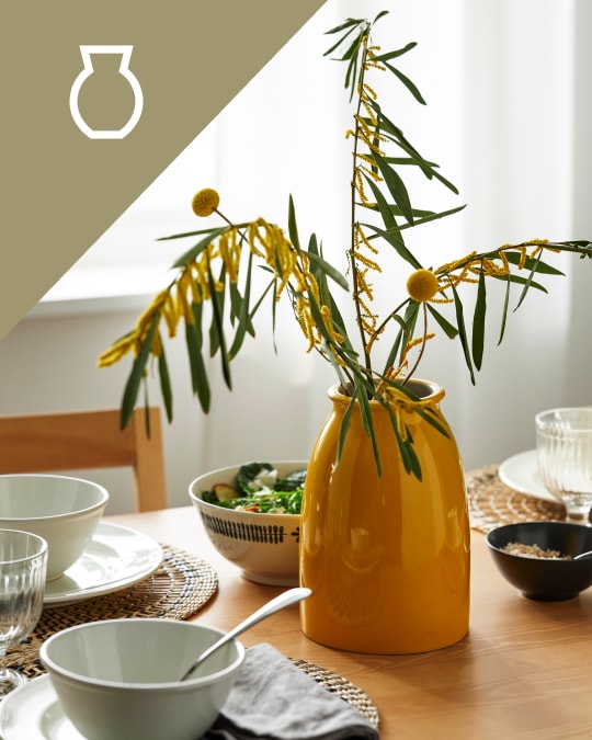 Una mesa con vajilla de desayuno y decorando la mesa un florero mostaza con plantas.