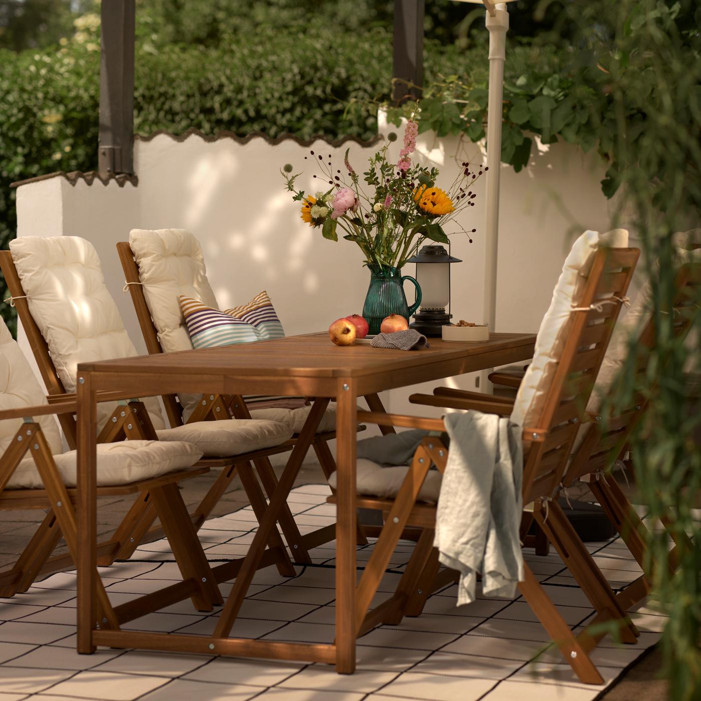 Una mesa de exterior de color marrón claro con flores en una jarra estampada/verde y seis sillas reclinables con cojines de asiento/respaldo beige.