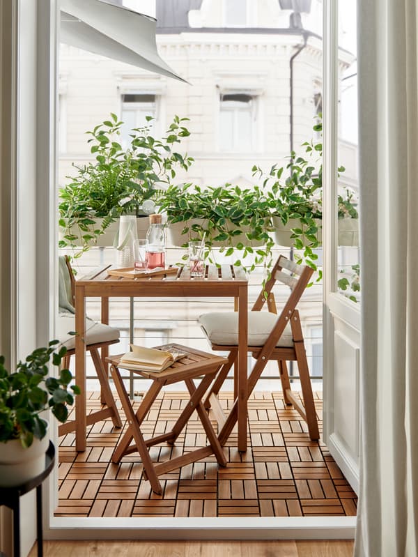 Una mesa de exterior NÄMMARÖ teñida de marrón claro, un taburete plegable y sillas plegables en un balcón rodeado de plantas frondosas.