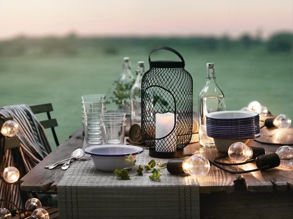 Una mesa de madera exterior con platos enlozados, un soporte para velas, vasos apilados, una botella KORKEN y una guirnalda SOLVINDEN con luces prendidas. 