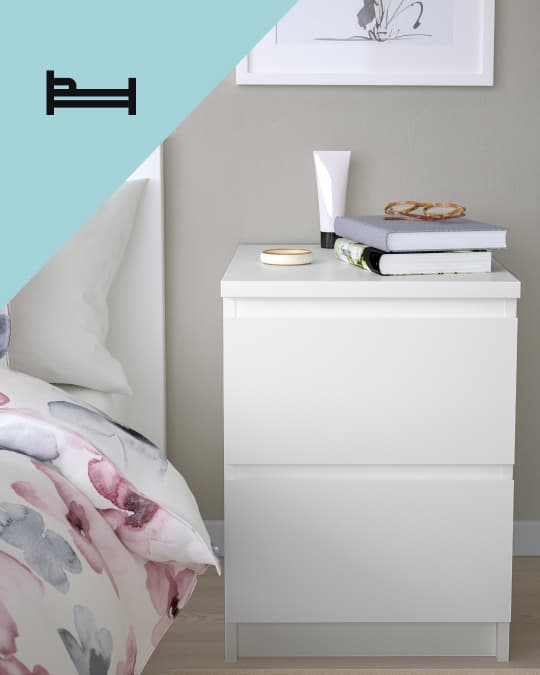 Una mesa de noche blanca co dos cajones al costado de una cama y en el costado superior izquierdo de la foto un ícono de cama.