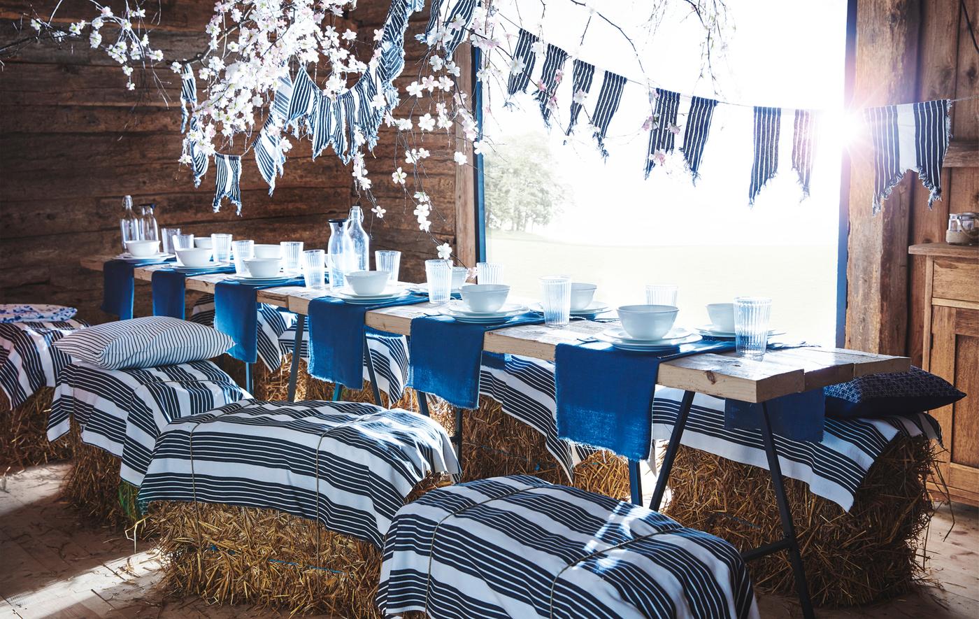 Una mesa larga en un cobertizo de madera con vajilla azul y blanca, y tela a rayas usada como banderín y atada a fardos de heno que se usan a modo de asientos.