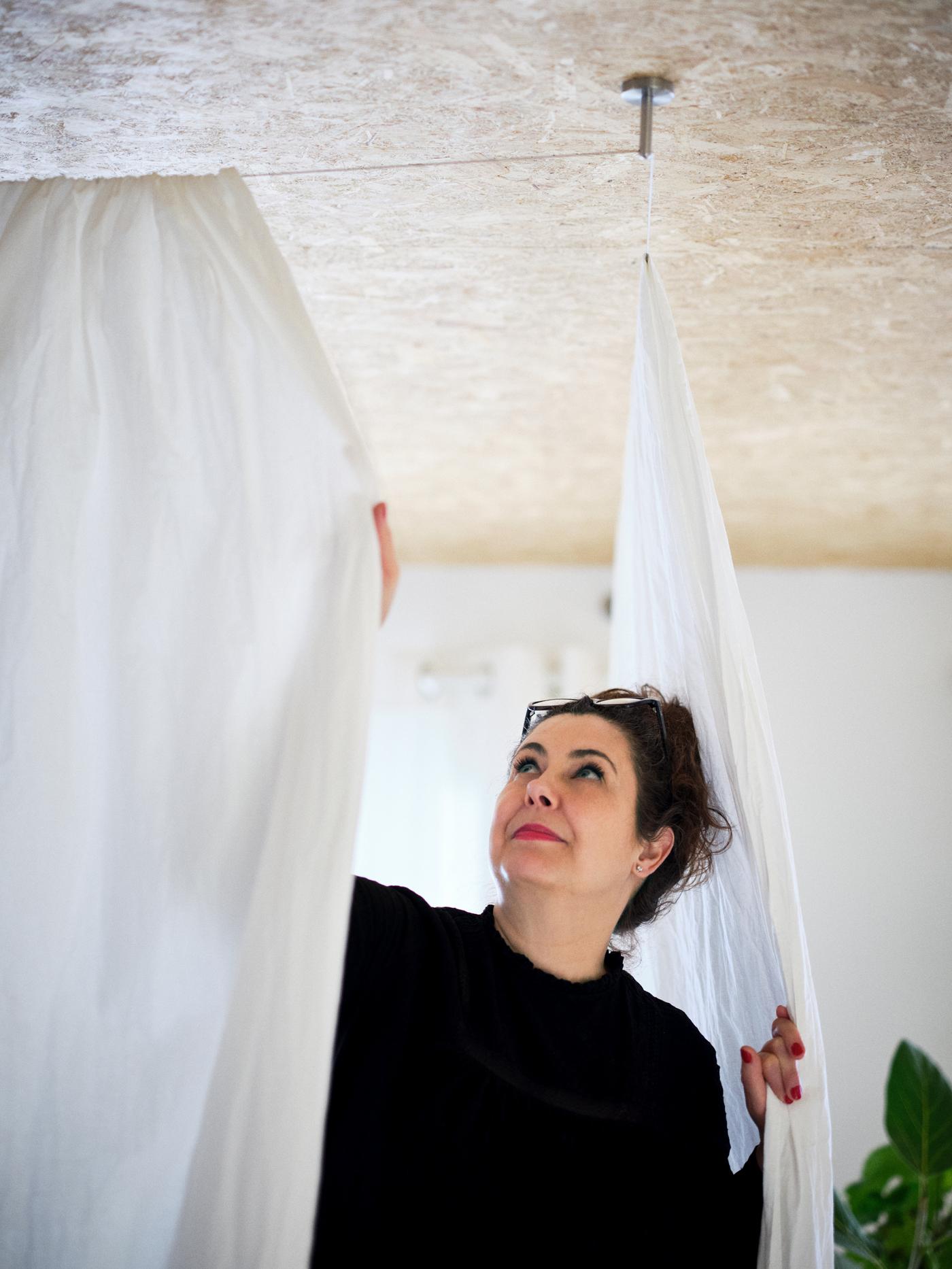 Una mujer cerrando dos cortinas blancas de algodón montadas en el techo y que dividen la habitación, con una gran planta en la esquina.